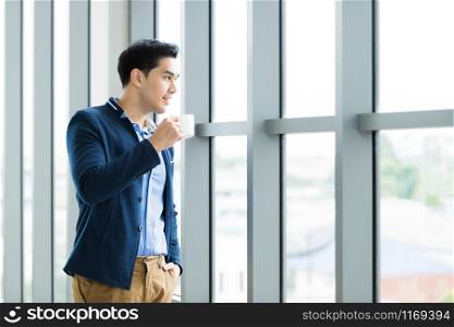 Portrait of Happy mood asian young businessman working holding a coffee cup wear a business suit of man in blue jacket and blue shirt looking at the window In the office room background