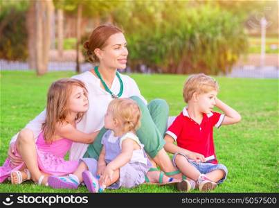 Portrait of happy mother with cute little babies sitting on fresh green grass field, looking away, having fun outdoors, enjoying parenthood, happiness and love concept
