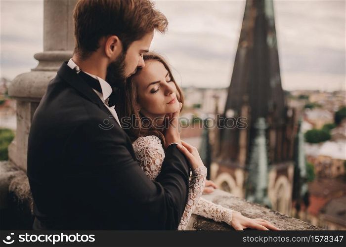 portrait of Happy newlywed. beautiful bride and stylish groom are hugging on the balcony of old gothic cathedral with panoramic city views.. portrait of Happy newlywed. beautiful bride and stylish groom are hugging on the balcony of old gothic cathedral with panoramic city views