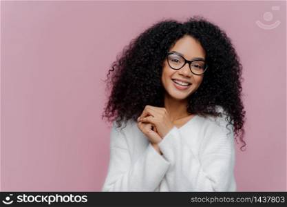 Portrait of happy positive African American woman with curly bushy hair, keeps hands together, has pleased facial expression, wears spectacles and white sweater, poses over purple background