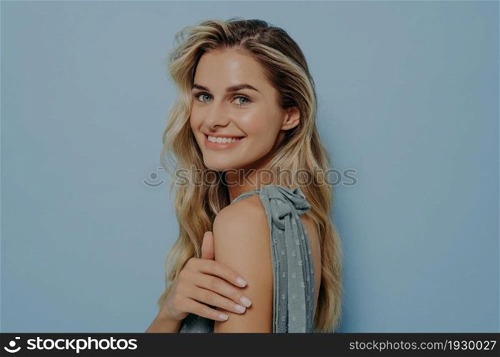 Portrait of happy positive smiling blonde woman wearing blue dress turning back to look over shoulder with elegant smile, holding her own hand while standing isolated over blue background. Happy positive smiling blonde woman looking over shoulder