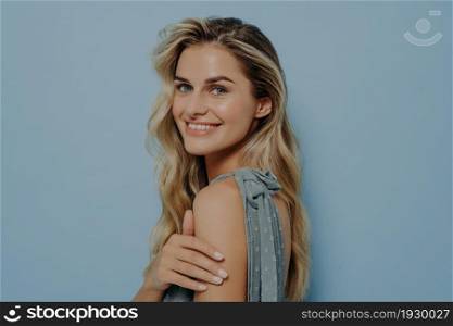 Portrait of happy positive smiling blonde woman wearing blue dress turning back to look over shoulder with elegant smile, holding her own hand while standing isolated over blue background. Happy positive smiling blonde woman looking over shoulder
