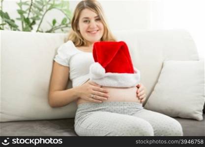 Portrait of happy pregnant woman posing with red Santa hat on belly