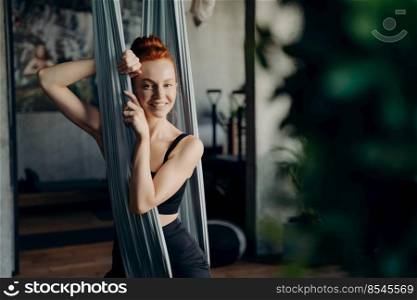 Portrait of happy red haired female fitness instructor in black sportswear sitting in hammock, waiting for clients to conduct anti gravity or aerial yoga class in studio, looking at camera with smile. Young red haired woman sitting in hammock while visiting aerial yoga class