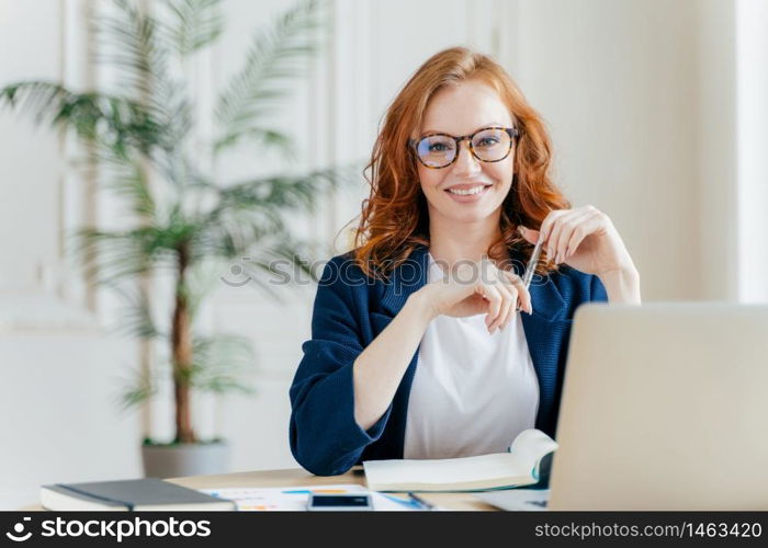 Portrait of happy redhaired woman employee in optical glasses, has satisfied expression, works with modern gadgets, waits for meeting with colleague, prepares accounting report, sits in own cabinet