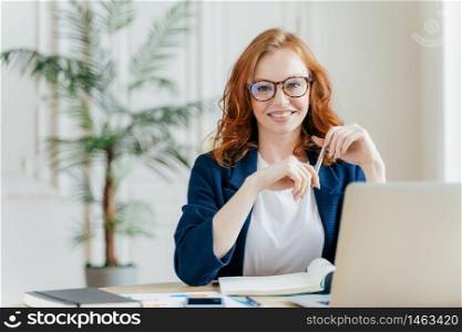 Portrait of happy redhaired woman employee in optical glasses, has satisfied expression, works with modern gadgets, waits for meeting with colleague, prepares accounting report, sits in own cabinet