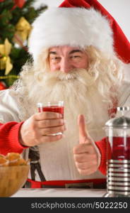 Portrait of happy Santa Claus at home looking at camera and thumbs up while eating cookies and drinking tea