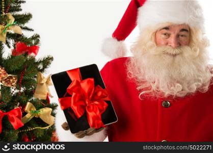 Portrait of happy Santa Claus holding gift Tablet computer in his hands with ribbon and looking at camera against Christmas tree