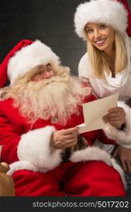 Portrait of happy Santa Claus sitting at his room at home with his woman helper near Christmas tree and big sack and reading Christmas letter or wish list