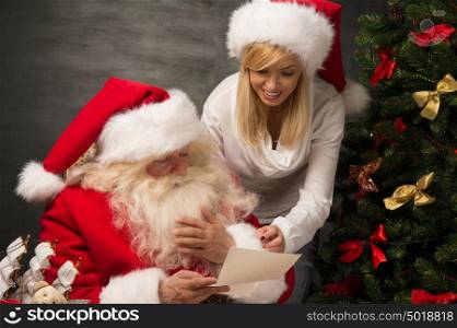 Portrait of happy Santa Claus sitting at his room at home with his woman helper near Christmas tree and big sack and reading Christmas letter or wish list