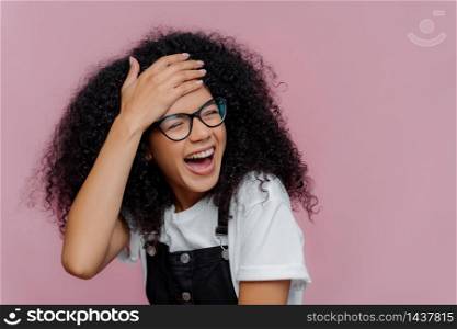 Portrait of happy satisfied African American woman touches forehead, feels energized and overemotive, wears optical glasses, has curly hair, laughs sinecerely, isolated on purple studio wall