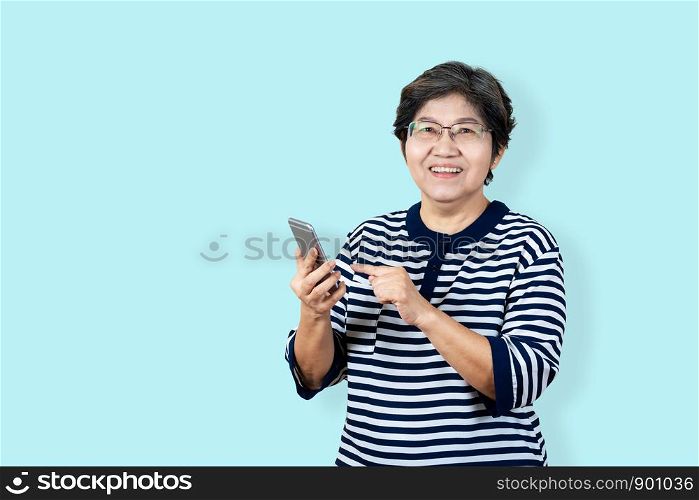 Portrait of happy senior asian woman holding or using smartphone and looking at camera on isolated background feeling positive enjoy and satisfaction. Older female lifestyle concept blue background.