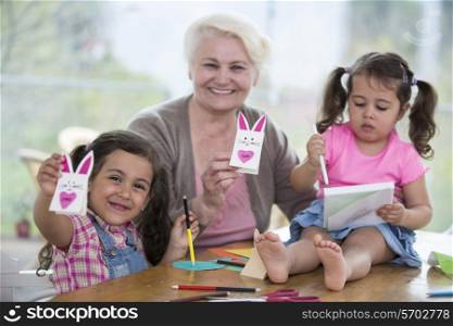 Portrait of happy senior woman showing craft rabbit while sitting with granddaughters at home
