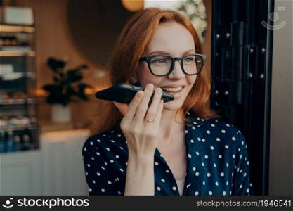 Portrait of happy smiling business woman in spectacles talks on cell phone on loudspeaker while leaving coffee shop in morning, redhead female record audio message while spending time outdoors. Happy smiling businesswoman talks on cell phone on loudspeaker while leaving coffee shop