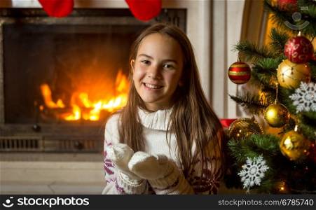 Portrait of happy smiling girl in sweater and gloves posing at burning fireplace and decorated Christmas tree