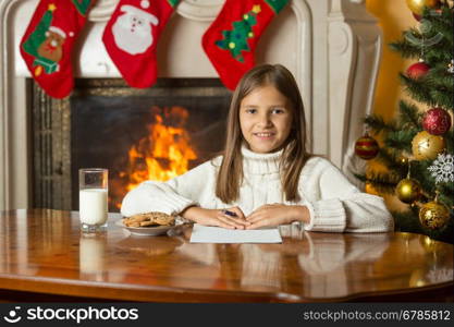 Portrait of happy smiling girl sitting by fireplace and writing letter to Santa Claus