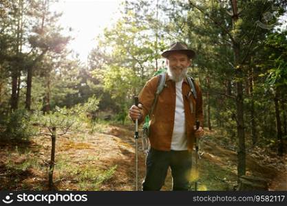Portrait of happy smiling mature man hiking in wood. Handsome senior male backpacker using poles sticks for active trekking walk along forest paths. Portrait of happy smiling mature man hiking in wood