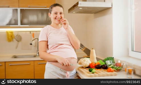 Portrait of happy smiling pregnant woman cooking on kitchen and wating fresh tasty vegetables. Concept of healthy lifestyle and nutrition during pregnancy.. Portrait of happy smiling pregnant woman cooking on kitchen and wating fresh tasty vegetables. Concept of healthy lifestyle and nutrition during pregnancy