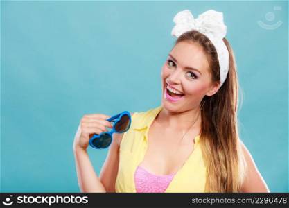 Portrait of happy smiling pretty pin up girl with hairband bow wearing heart shaped sunglasses. Attractive gorgeous young retro woman posing in studio on blue background.