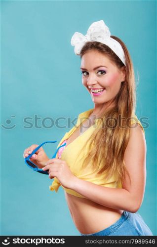 Portrait of happy smiling pretty pin up girl with hairband bow wearing heart shaped sunglasses. Attractive gorgeous young retro woman posing in studio on blue background.