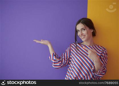 Portrait of happy smiling young beautiful woman in a presenting gesture with open palm isolated on purple and yellow background. Female model in modern fashionable clothes posing in the studio