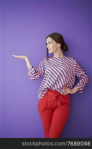 Portrait of happy smiling young beautiful woman in a presenting gesture with open palm isolated on purple background. Female model in modern fashionable clothes posing in the studio
