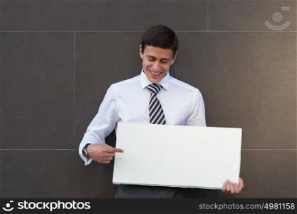 Portrait of happy smiling young businessman showing blank signboard, with copyspace area for text or slogan, against grey wall background outdoors