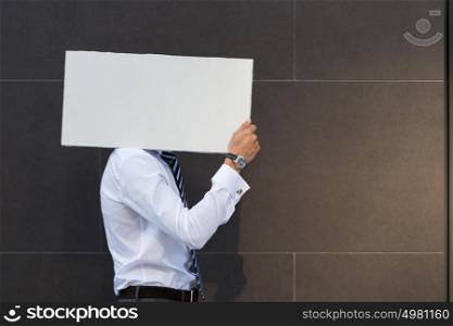 Portrait of happy smiling young businessman showing blank signboard, with copyspace area for text or slogan, against grey wall background outdoors
