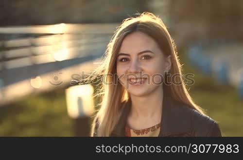 Portrait of happy smiling young caucasian woman with long blonde hair and perfect skin looking at camera with radiant smile at sunset in backlight. Positive emotion and facial expression concept.
