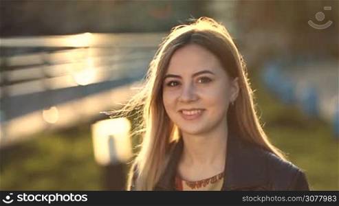 Portrait of happy smiling young caucasian woman with long blonde hair and perfect skin looking at camera with radiant smile at sunset in backlight. Positive emotion and facial expression concept.
