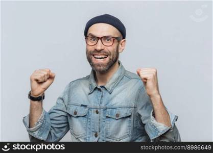 Portrait of happy successful man rejoices his triumph, clenches teeth, has overjoyed expression, isolated over grey background. Handsome fashionable male celebrates his victory. Achievement concept