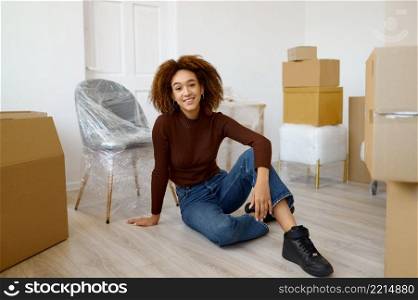 Portrait of happy tired young woman sitting on floor among boxes with beginnings. Home moving. Woman sitting among carton boxes with beginnings