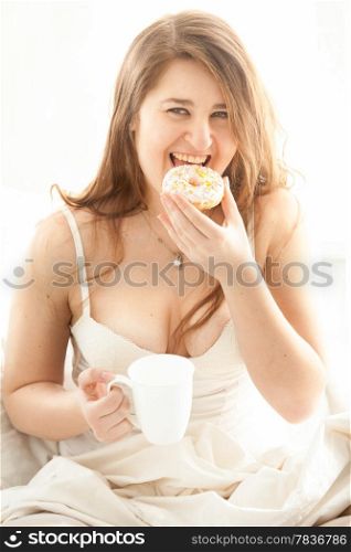 Portrait of happy woman eating donut and drinking coffee in bed