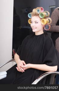 Portrait of happy woman in beauty salon. Cheerful blond girl with hair curlers rollers by hairdresser. Hairstyle.