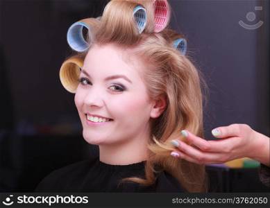 Portrait of happy woman in beauty salon. Smiling blond girl with hair curlers rollers by hairdresser. Hairstyle.