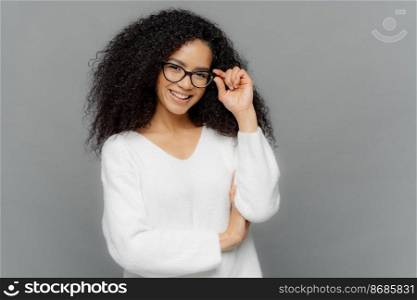 Portrait of happy woman with curly hair, kepes hands on frame of glasses, has gentle smile, wears white jumper, isolated over grey background. Copy space for your advert. Delighted student indoor