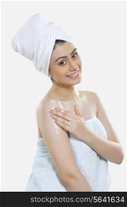Portrait of happy woman wrapped in towel applying moisturizer on arm against white background