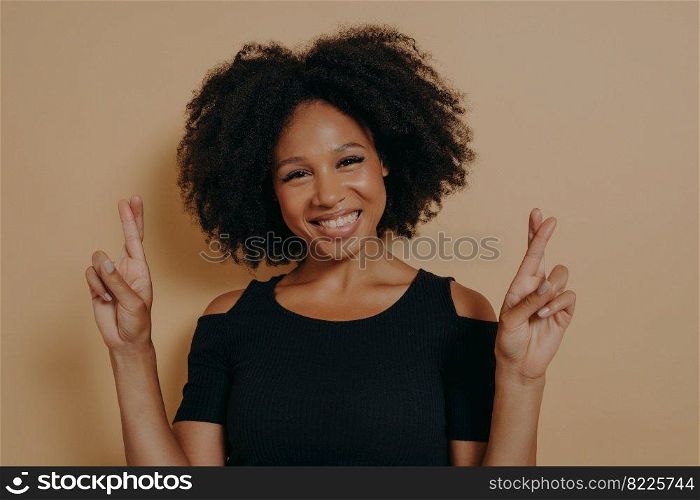 Portrait of happy young african woman standing isolated over dark beige studio background with copy space, holding fingers crossed for good luck broadly smiling. Positive emotions and body language. Portrait of happy young african woman standing isolated over dark beige background with copy space