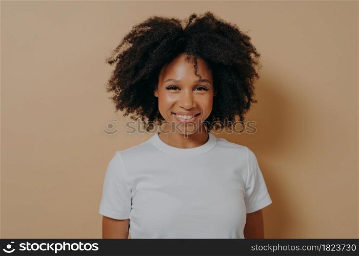 Portrait of happy young african woman with broad shining smile dressed in white tshirt posing alone against beige background, positive mixed race female being in high spirit. Positive people concept. Portrait of cheerful dark skinned woman expressing happiness while standing on beige background