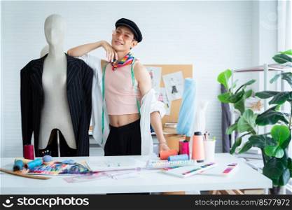 Portrait of happy young Asian LGBTQ gay dressmaker standing confident pose with mannequin behind working desk, sketch of fashion dress design, tailor tools in studio, small business owner concept