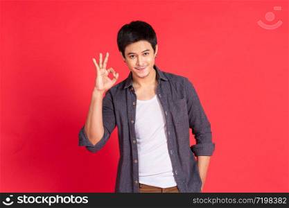 Portrait of happy young asian man dress in casual informal smiling and posing with cheerful and okey hand sign isolated on red background.