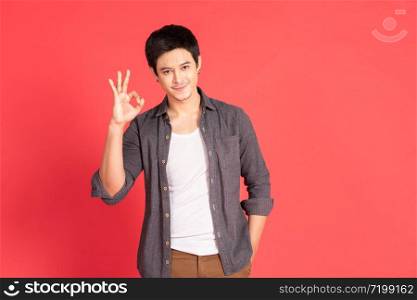 Portrait of happy young asian man dress in casual informal smiling and posing with cheerful and okey hand sign isolated on red background.