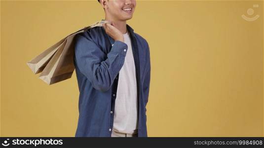 portrait of happy young asian man dressed casually holding shopping bags isolated on yellow studio background