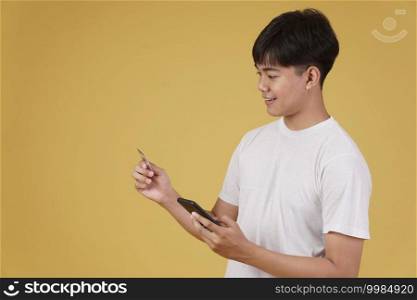 portrait of happy young asian man dressed casually holding smartphone and credit card for shopping online isolated on yellow studio background