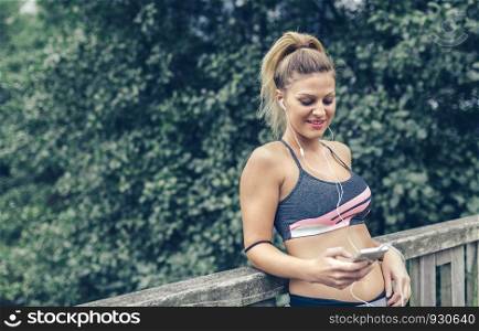 Portrait of happy young blonde woman in sports wear with earphones looking her smartphone after training outdoors. Woman in sports wear with earphones looking smartphone