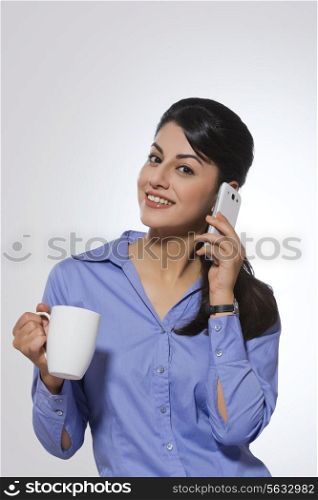 Portrait of happy young businesswoman with coffee mug answering phone over gray background