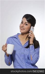 Portrait of happy young businesswoman with coffee mug answering phone over gray background