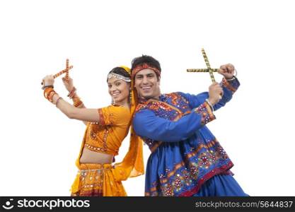 Portrait of happy young couple in traditional wear standing back to back while performing Dandiya Raas over white background