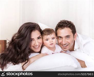 Portrait of happy young family at home in the morning, lying down in the bed, mother and father having fun with little daughter, love concept