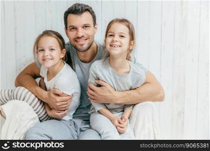 Portrait of happy young father embraces his two daughters, loves them very much, pose together at home. Single dad with beautiful femle children have cheerful expressions. Fatherhood concept